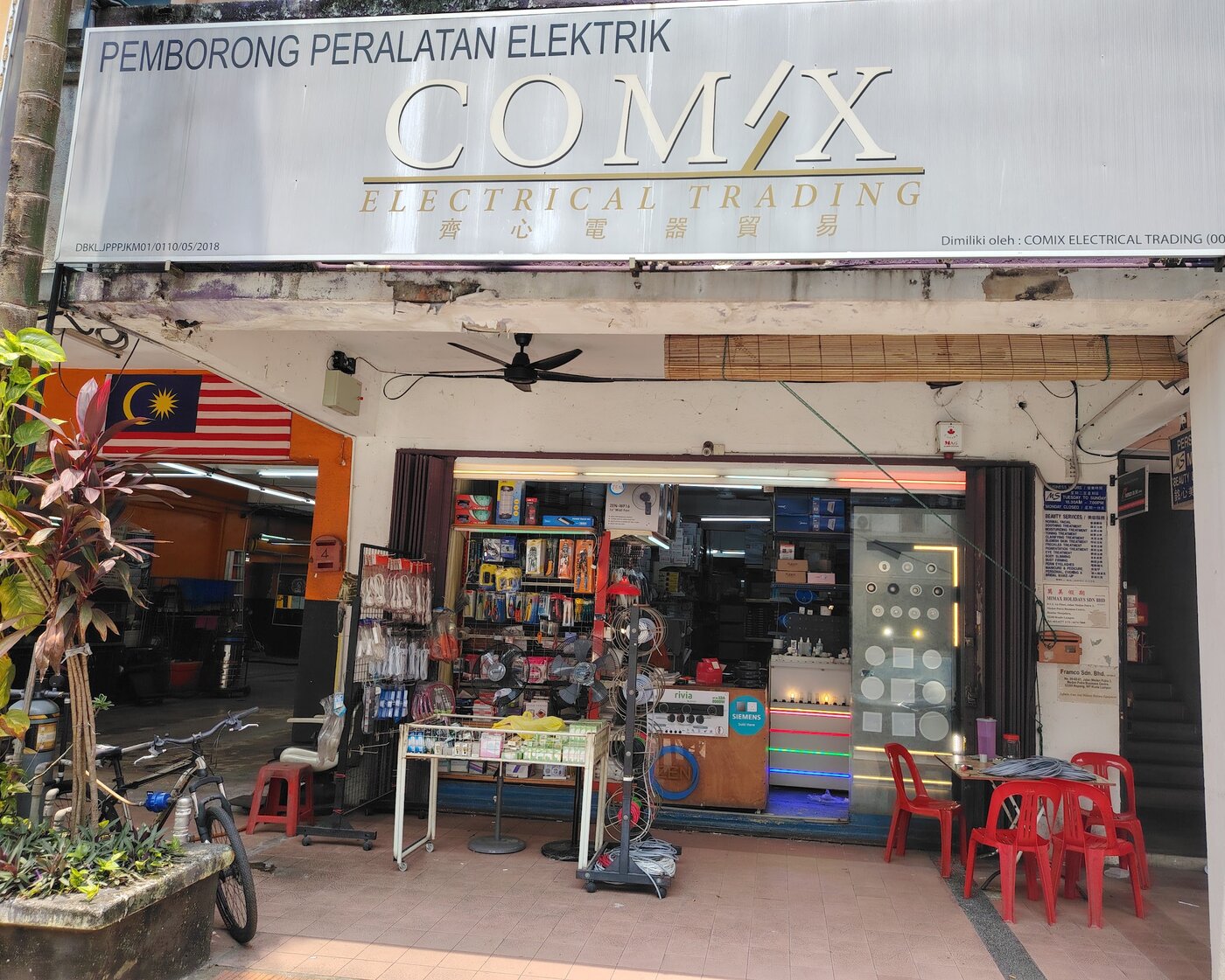 Shopfront with a grey signboard reading Pemborong Peralatan Elektrik, Comix Electrical Trading, a rack of cable reels, hanging extension sockets and a lit lighting display