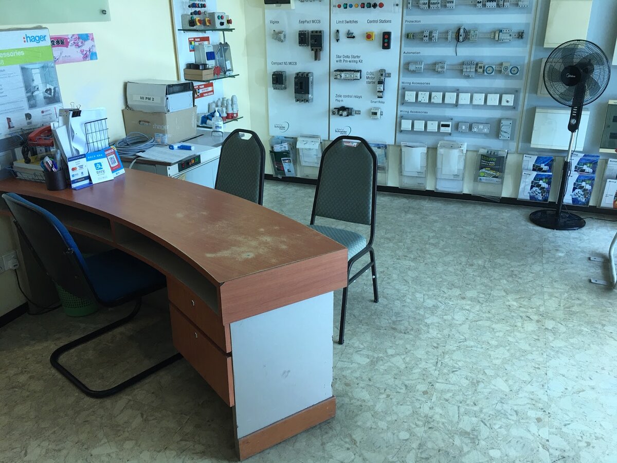 Office counter with two chairs in front of wall-mounted display boards of breakers, push buttons and enclosures, one headed Hager