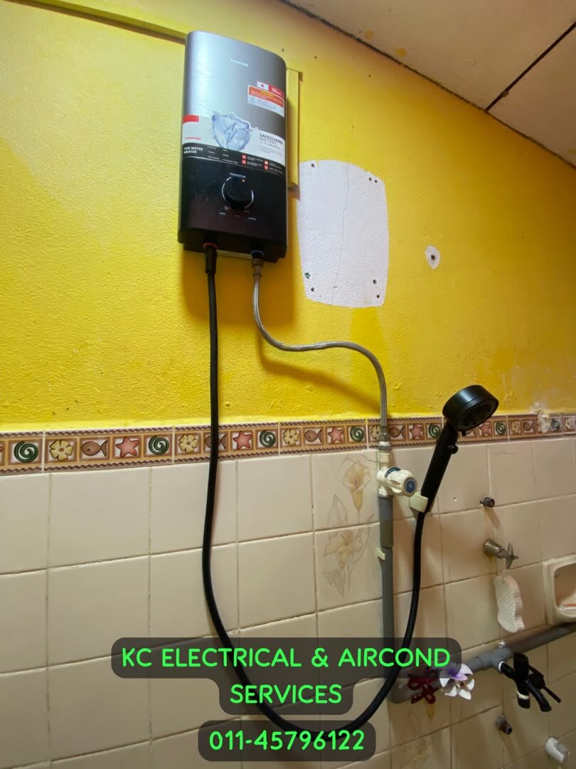 Grey and black instant water heater on a yellow bathroom wall with a hand shower and hose, watermarked with KC Electrical & Aircond Services and 011-45796122