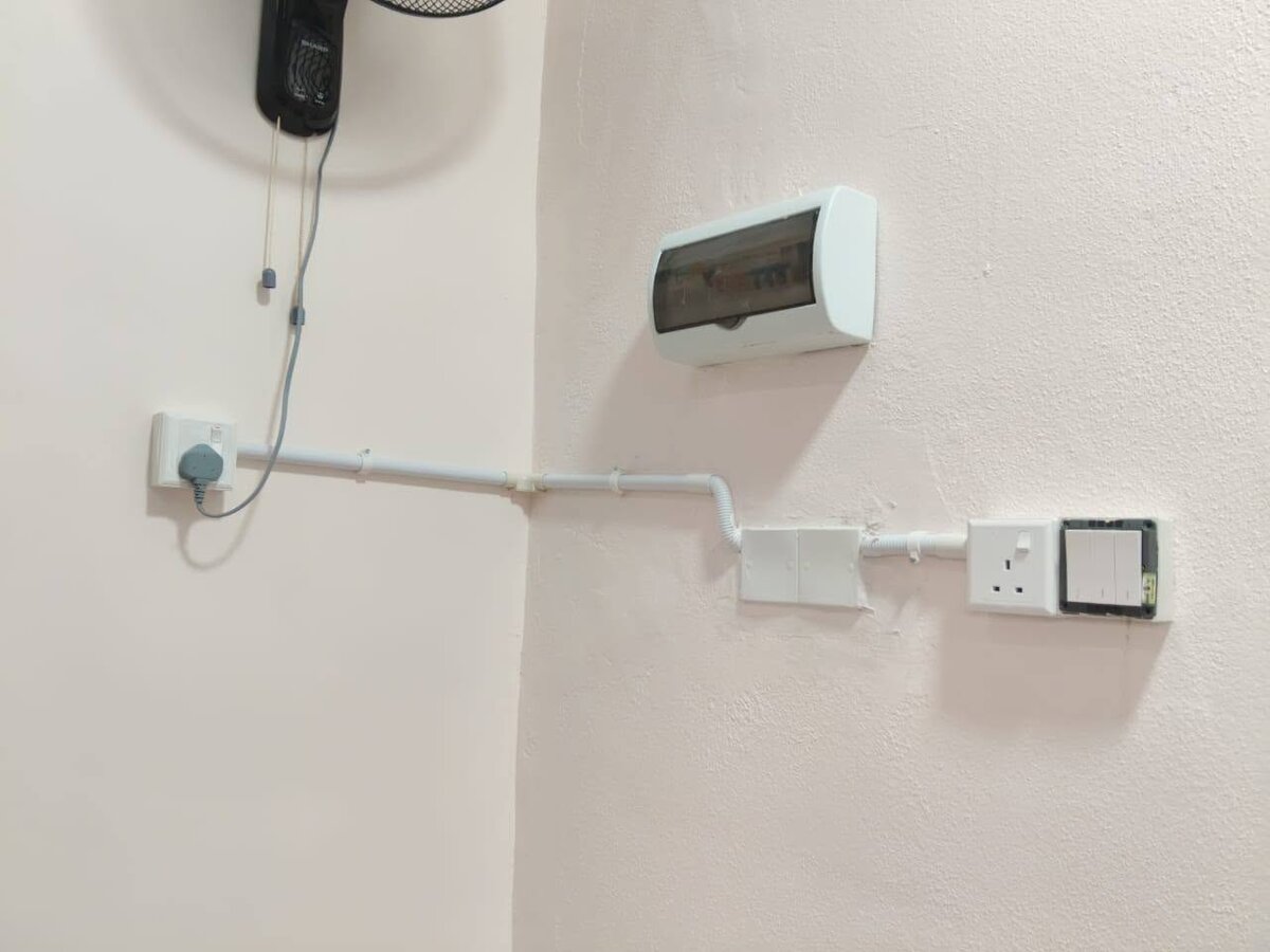 Pink wall with a socket on the left, white PVC conduit running around the corner to a blank box and a socket-and-switch plate, and a small white DB with a smoked cover above