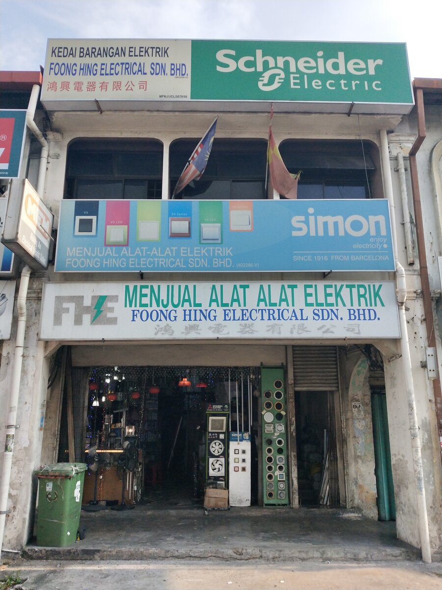 Two-storey shophouse with three signboards: Foong Hing Electrical Sdn Bhd with a green Schneider Electric panel, a blue Simon switch banner, and a white FHE sign; ventilating-fan and switch display stands at the open shopfront