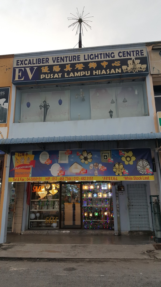 Two-storey shop lot with a navy signboard reading Excaliber Venture Lighting Centre, EV Pusat Lampu Hiasan, a promotional banner of downlights and ceiling lights with phone numbers, and a lit glass shopfront of chandeliers below