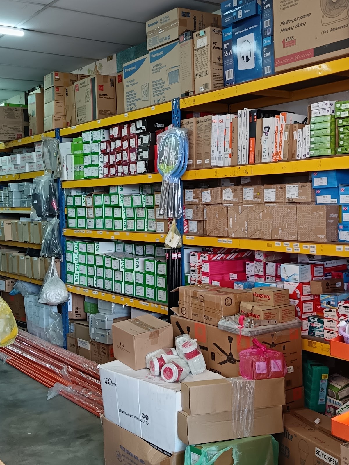 Yellow metal shelving stacked with green switch boxes, Hager cartons and fan boxes, with orange pipes and cartons on the floor