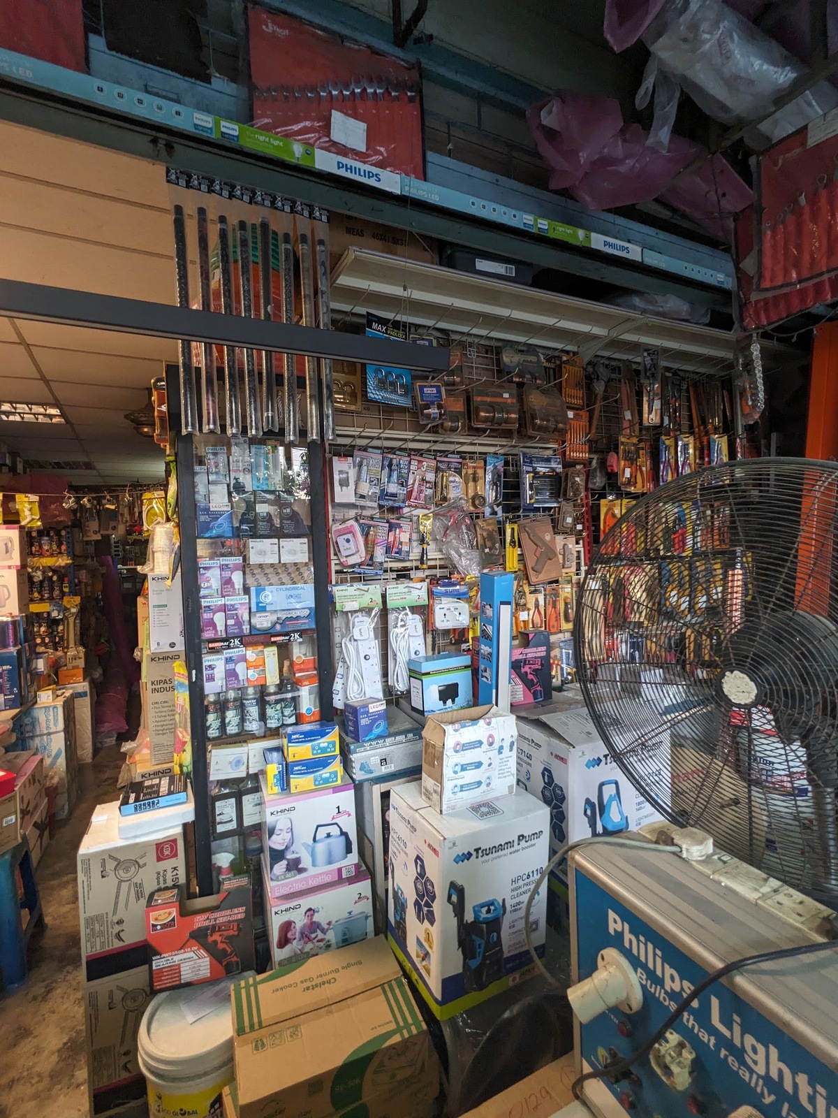 Crowded shop interior with Philips lamp boxes on a shelf, hanging extension sockets, padlocks and tools on pegboards, and boxed appliances and a stand fan in the foreground
