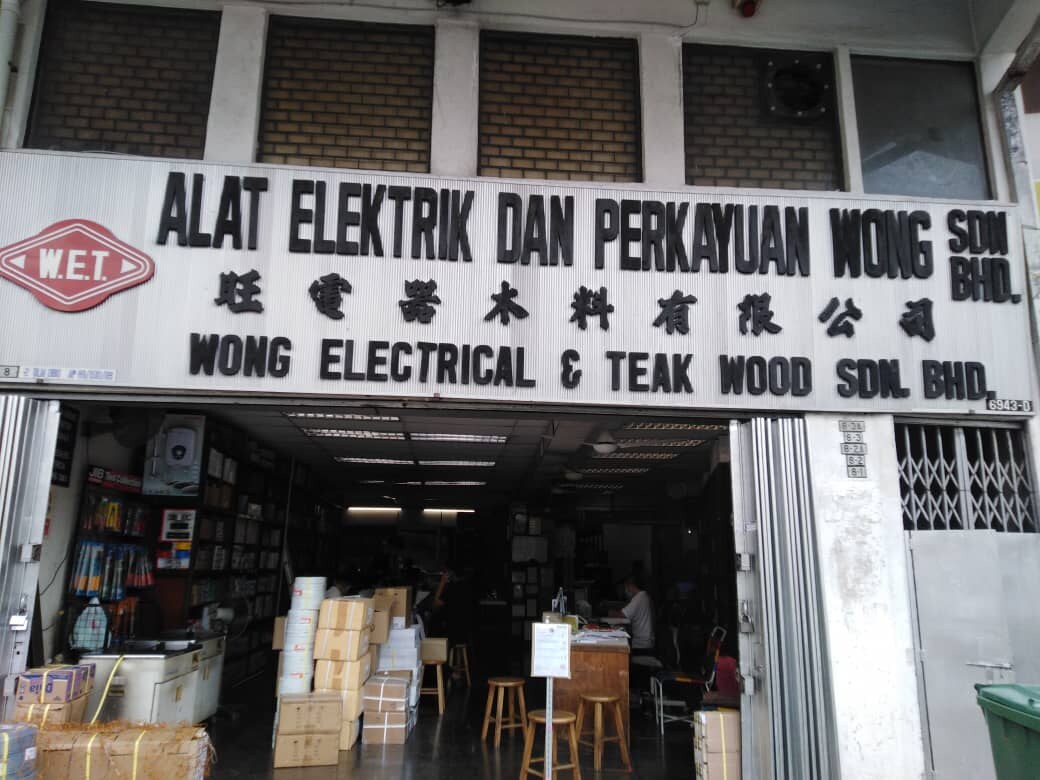 White fascia with black lettering reading Alat Elektrik dan Perkayuan Wong Sdn Bhd and Wong Electrical & Teak Wood Sdn Bhd above an open shop with boxed stock and stools