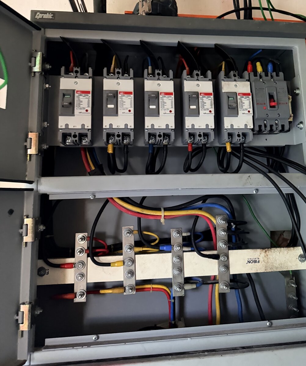 Grey metal distribution board, doors open, with a row of six moulded-case circuit breakers at the top and red, yellow, blue and black cables connected to four busbar links below