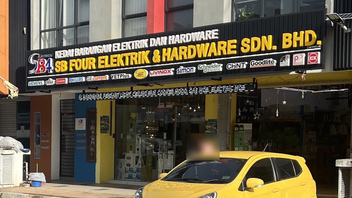 Black fascia reading Kedai Barangan Elektrik dan Hardware, SB Four Elektrik & Hardware Sdn. Bhd., over a two-unit shopfront with a yellow car parked in front
