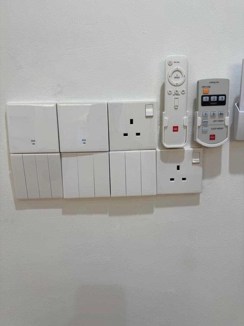 White wall plate with rows of rocker switches and two 13A sockets, with a fan remote and an aircond remote in holders beside them