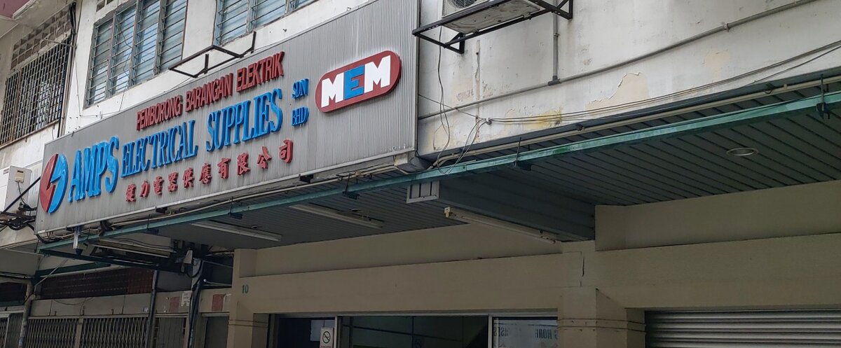 Silver signboard with blue AMPS Electrical Supplies lettering, red Malay and Chinese text and a red MEM logo above a shophouse walkway