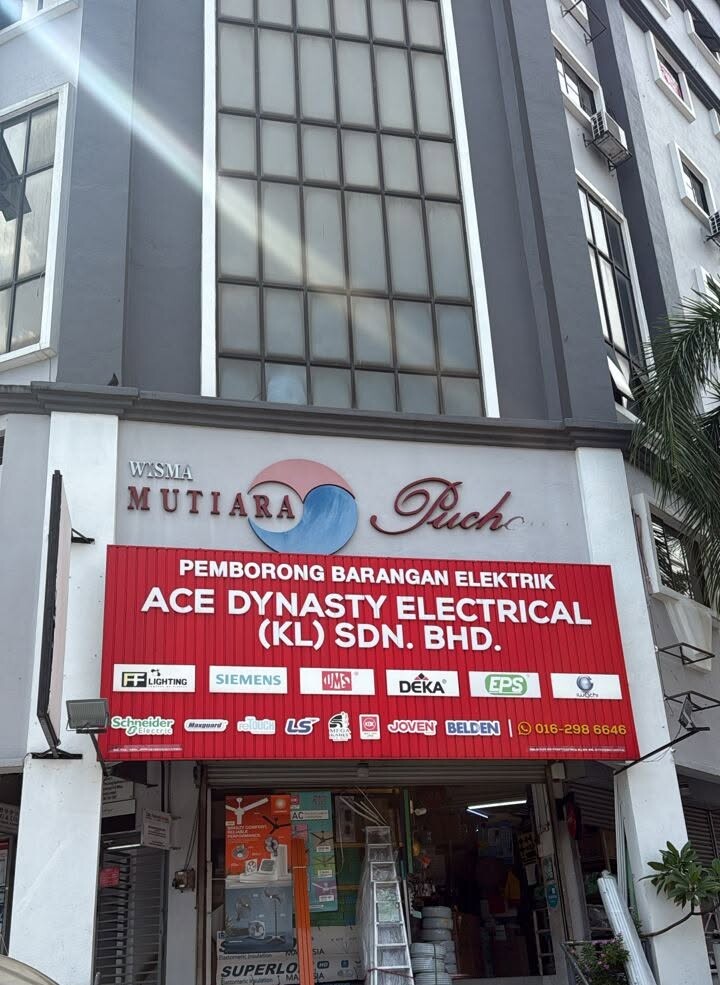 Red signboard reading Ace Dynasty Electrical (KL) Sdn Bhd with rows of brand logos under the Wisma Mutiara building sign, with stock and a ladder in the doorway