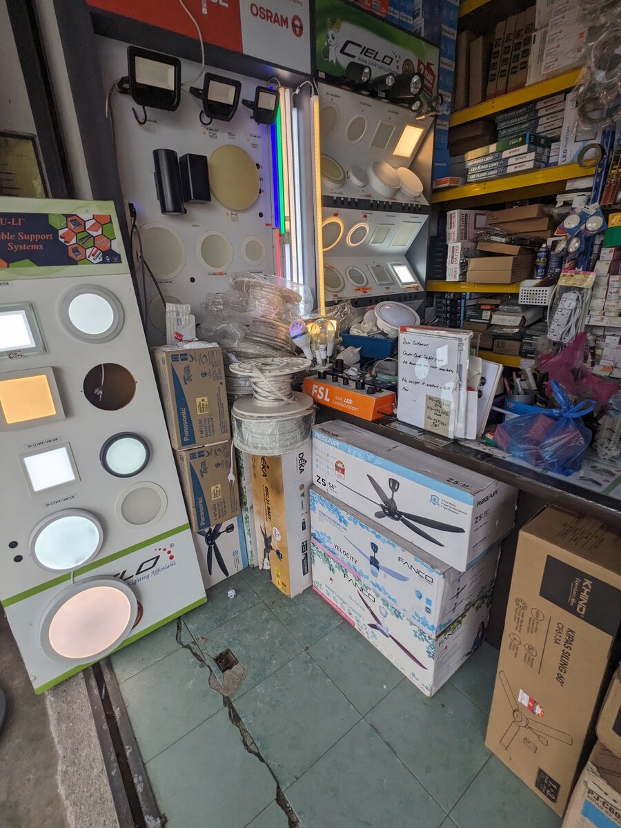Shop interior with a wall of sample downlights and floodlights, reels of LED strip, boxed ceiling fans on the floor and shelves of electrical parts