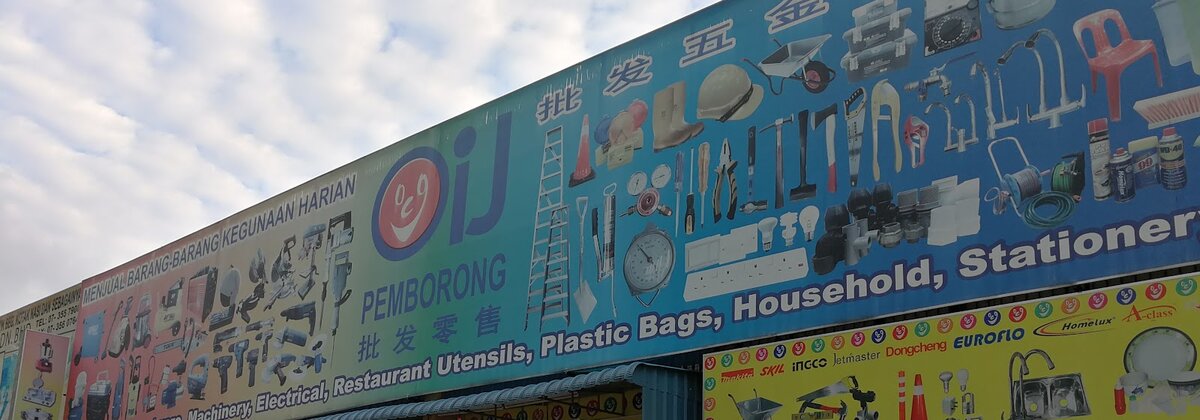 Long blue banner reading "Pemborong" with pictures of tools, ladders, taps, light bulbs, switches and trailing sockets, and a yellow board of brand logos below