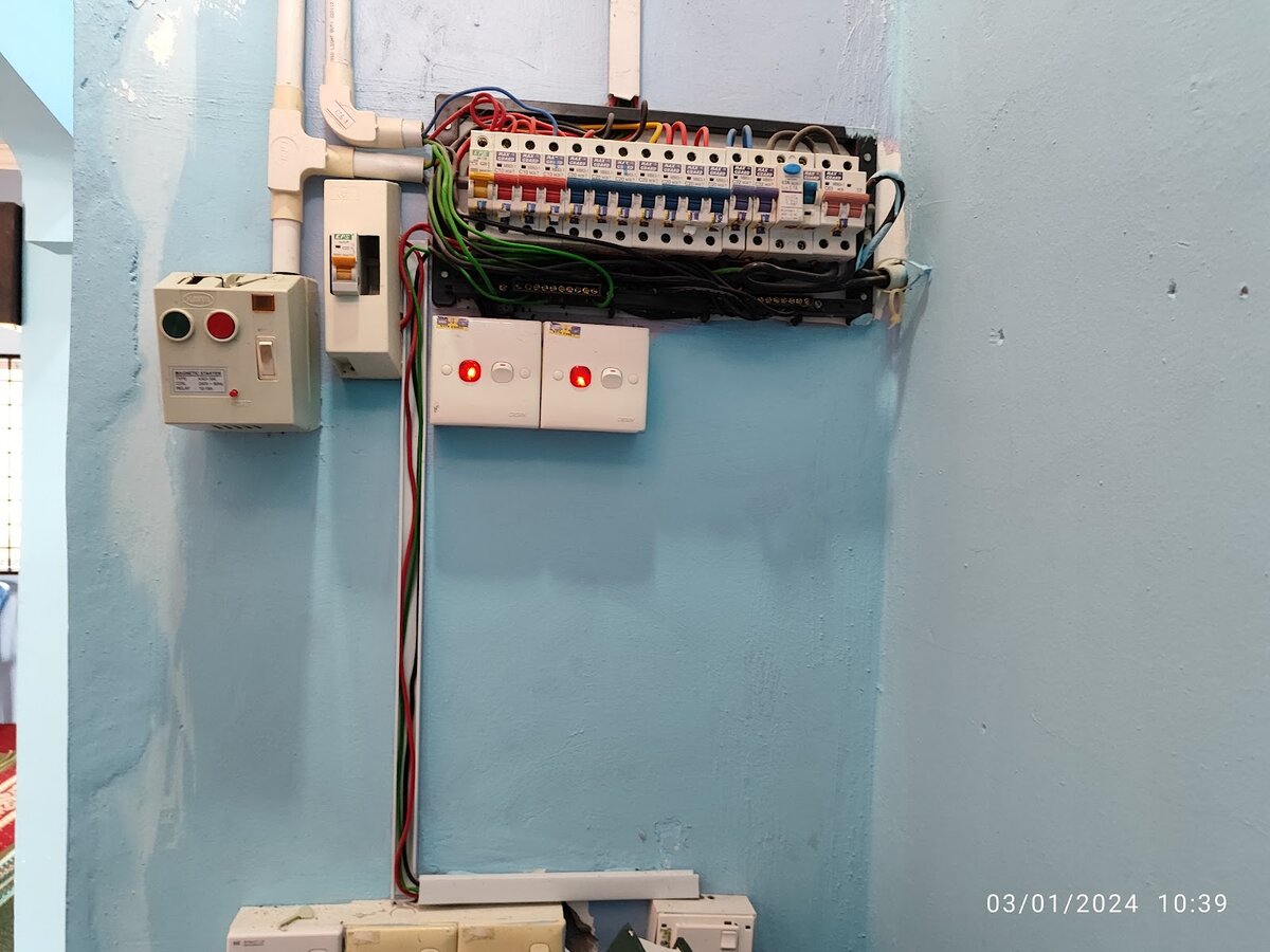 Wall-mounted distribution board with a row of Maxguard miniature circuit breakers and an RCCB, beside a grey magnetic starter with red and green buttons and two light switches with red neon indicators, on a pale blue wall