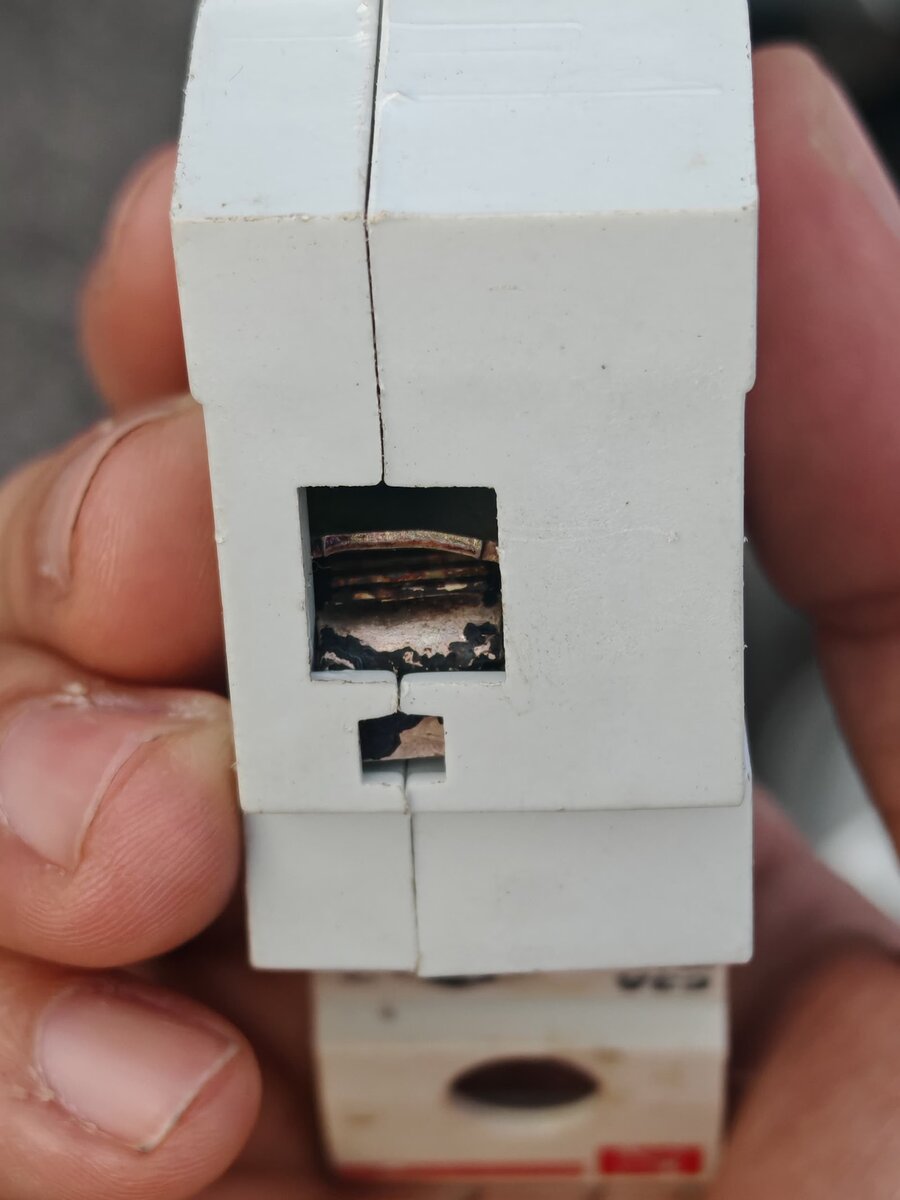 A hand holding a white modular breaker; through its cable-entry opening the terminal clamp is blackened and charred