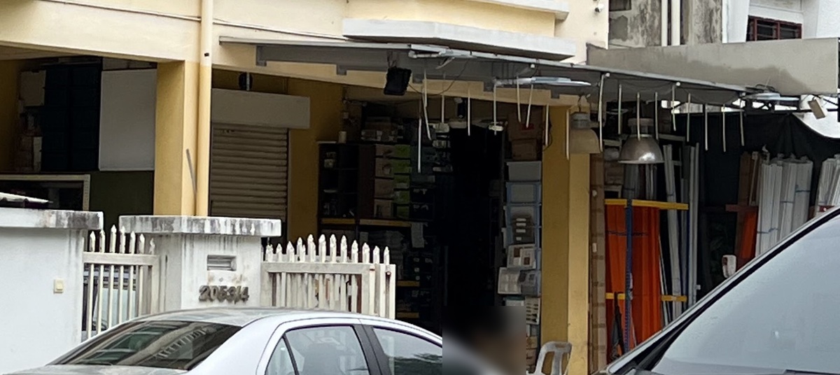 Ground floor of a yellow three-storey building behind a white picket gate, with shelves of boxes, switch display boards and hanging lamps under an awning