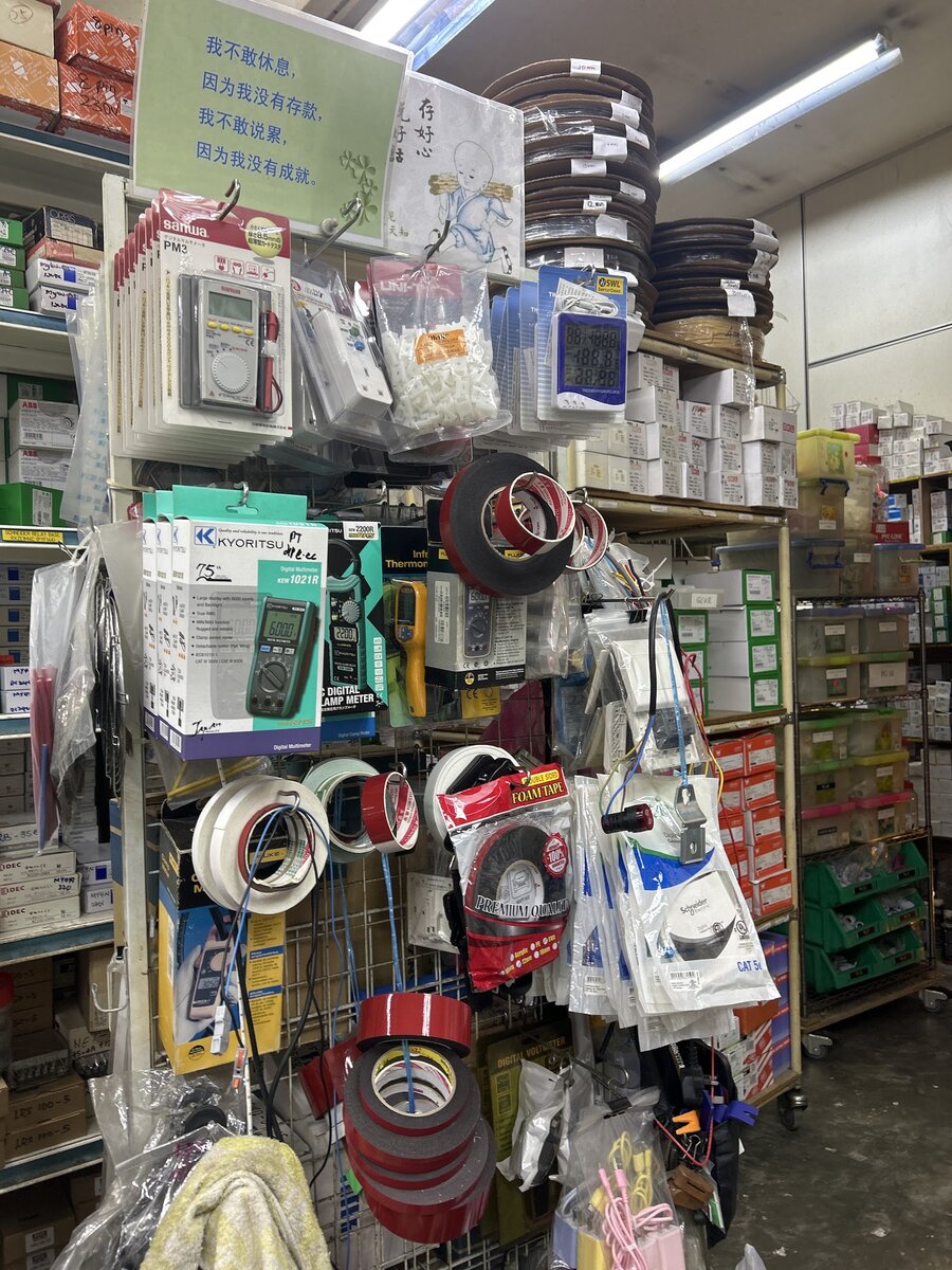 Shop aisle with hanging racks of electrical tapes, cable ties and boxed test meters, shelves of boxed parts and cable reels above