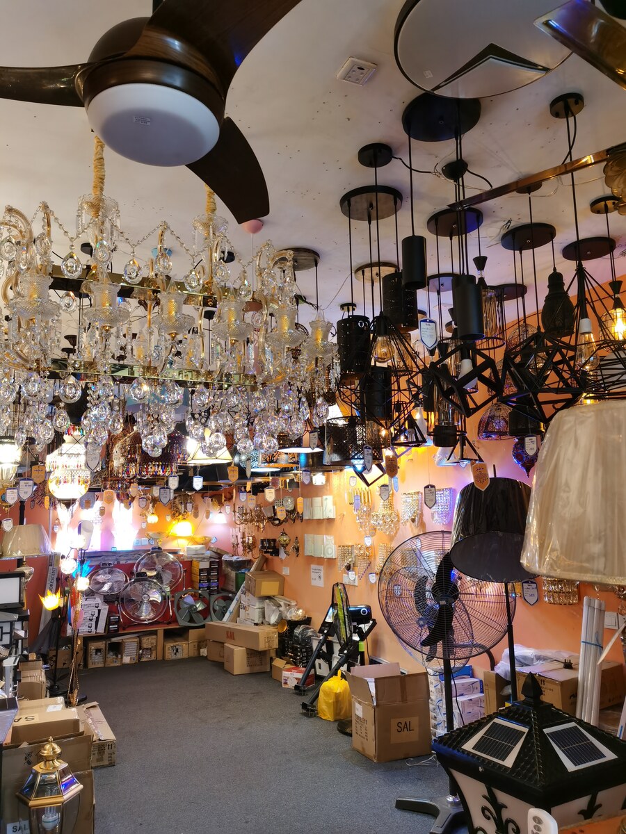 Shop interior crowded with chandeliers, black pendant lights and wall lamps, with stand fans and cartons on a grey carpet