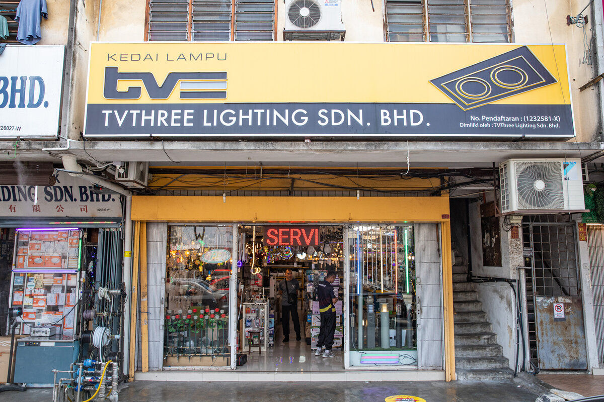 Yellow and grey signboard reading Kedai Lampu and TVThree Lighting Sdn. Bhd. over a glass shopfront with pendant lamps, LED strips and boxed lights inside