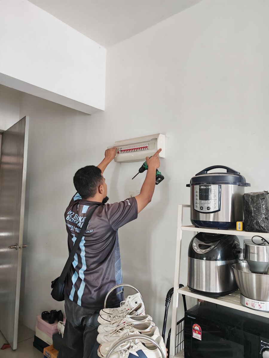 Technician in a dark company shirt drilling at a wall-mounted distribution board in a white room, beside a shelf of kitchen appliances and a pile of shoes