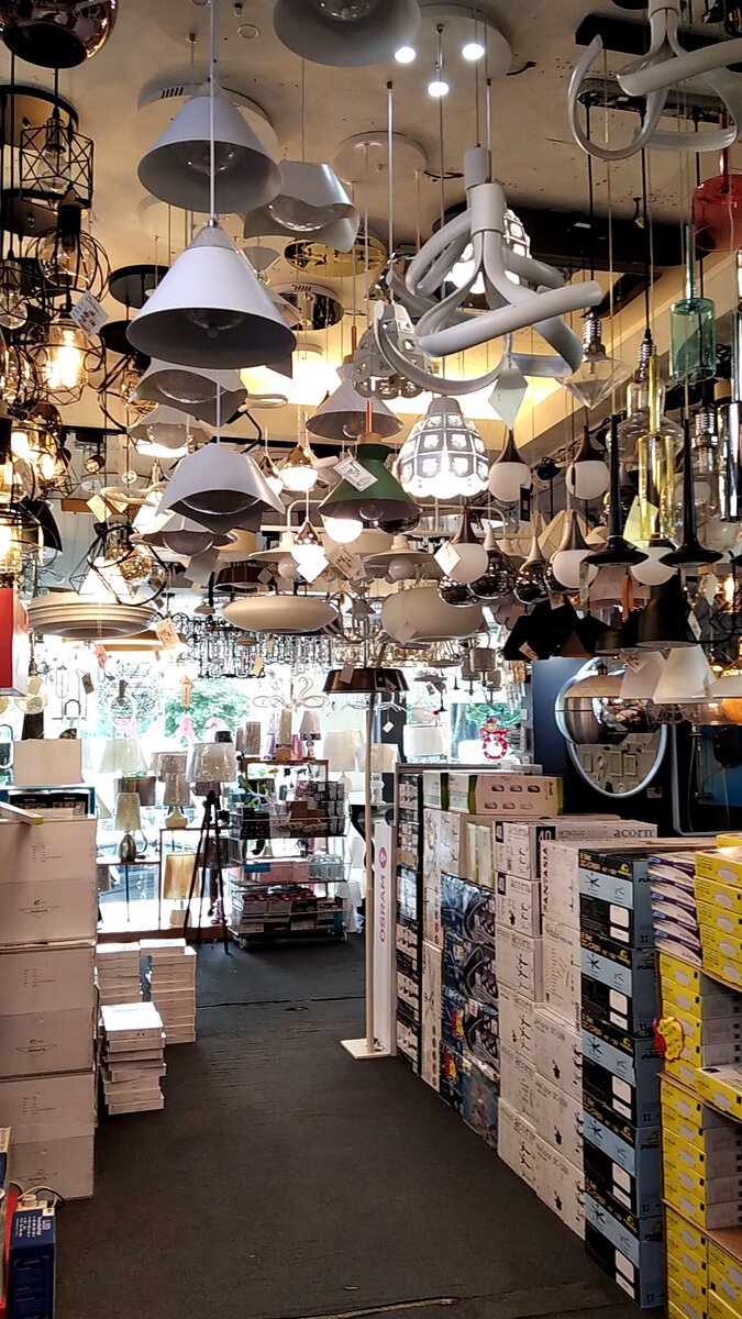 Narrow shop aisle under dozens of hanging pendant lamps, with stacked fan and lighting boxes on the right