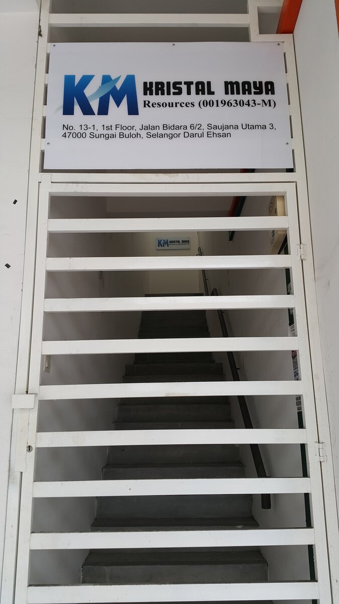 White signboard with a blue KM logo reading Kristal Maya Resources (001963043-M) and the Jalan Bidara 6/2, Saujana Utama 3 address, above a white stair gate