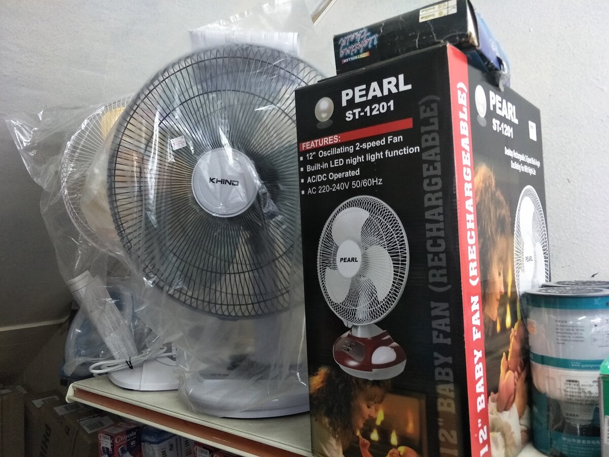 A stand fan in plastic wrap beside a boxed Pearl 12-inch rechargeable fan on a shelf