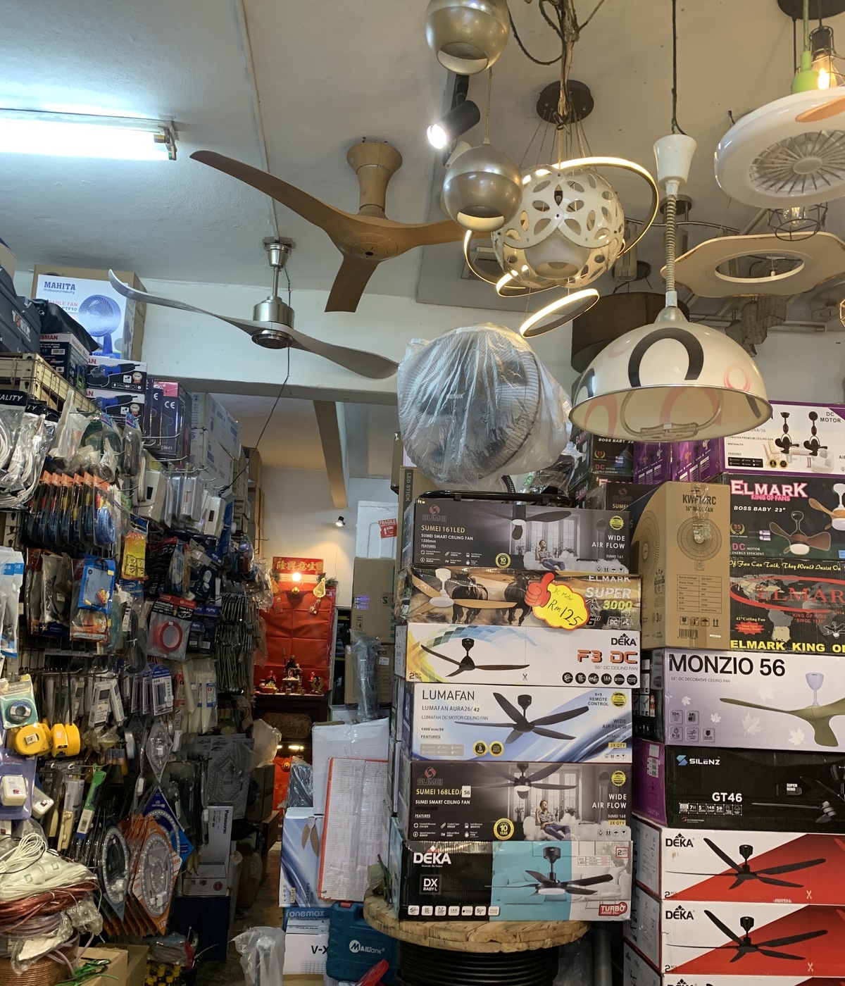 Crowded electrical shop with ceiling fans and pendant lamps hanging from the ceiling, towers of boxed ceiling fans and a wall of hanging packaged accessories