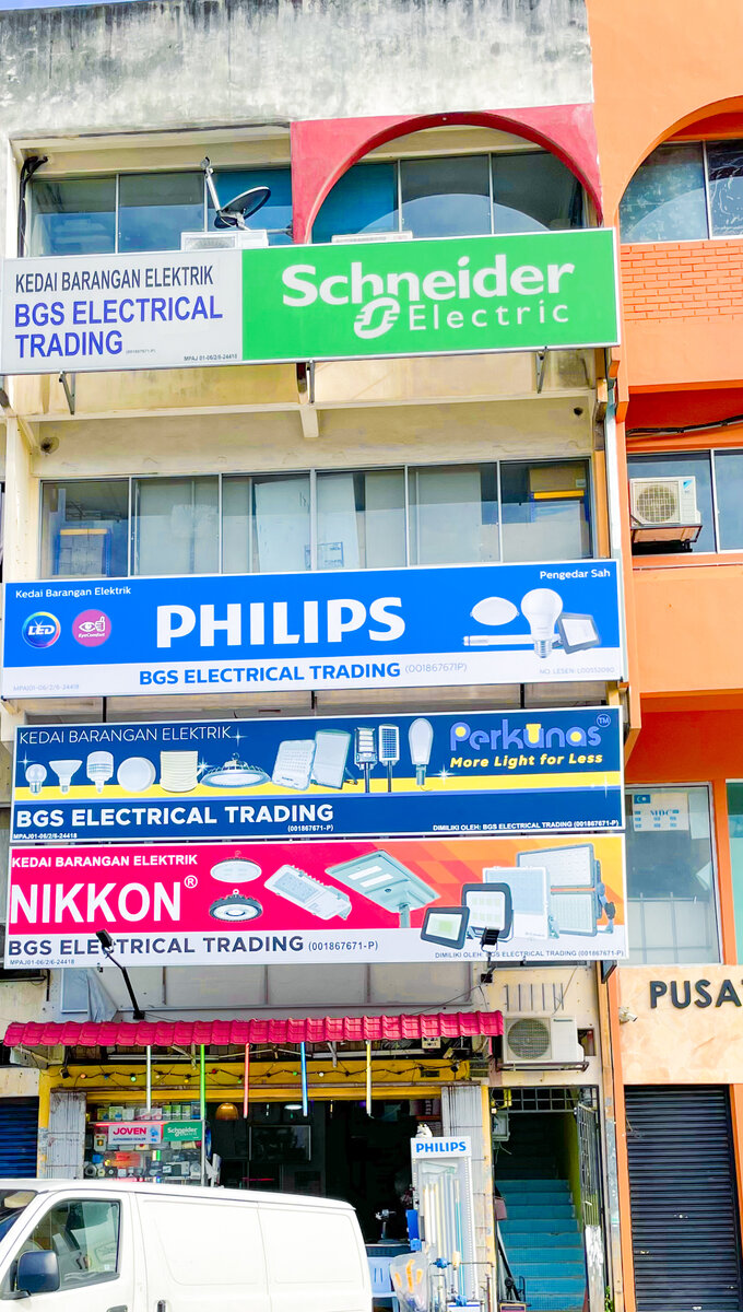 Four stacked shop signboards on a shophouse, green Schneider Electric, blue Philips, dark blue Perkunas and red Nikkon, each with BGS Electrical Trading, above an open shopfront with hanging coloured lights and a Philips display stand