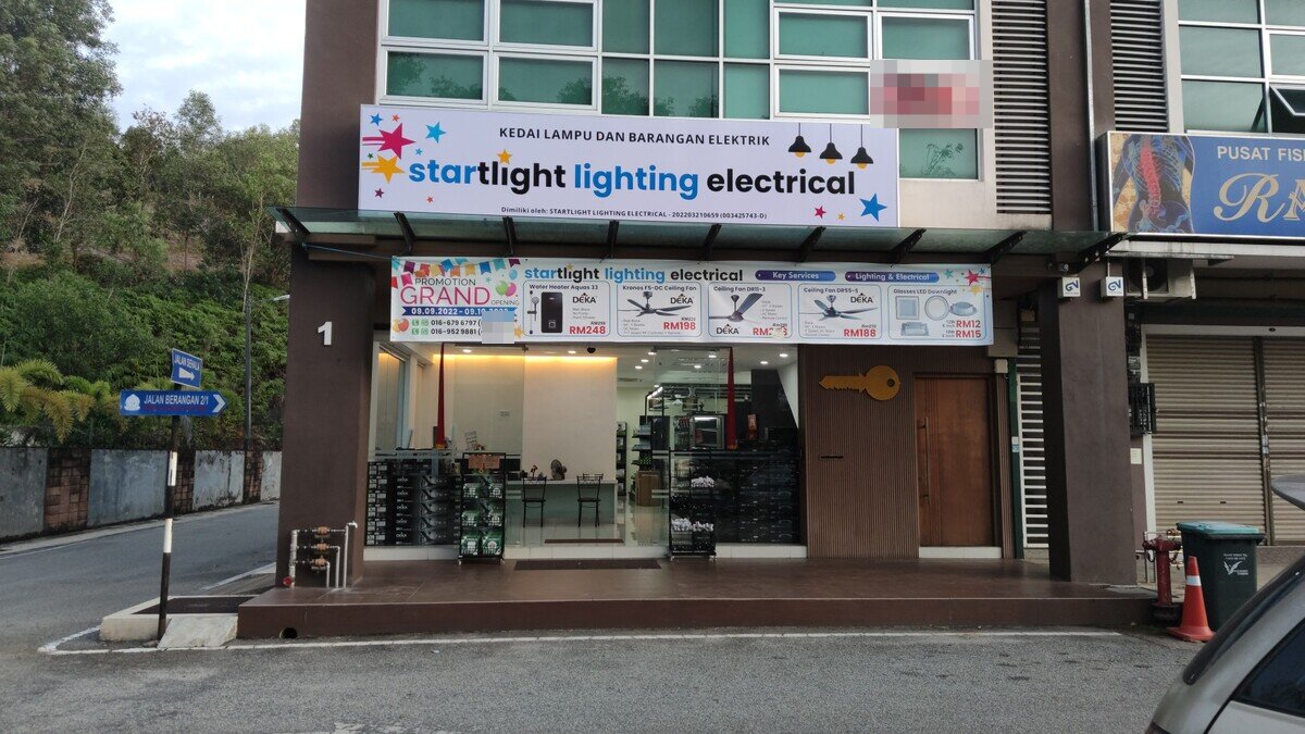 Corner shophouse (upper-floor sign and names on the banner blurred) with a white Startlight Lighting Electrical signboard, a grand-opening promotion banner and a glass shopfront with stacked boxes inside