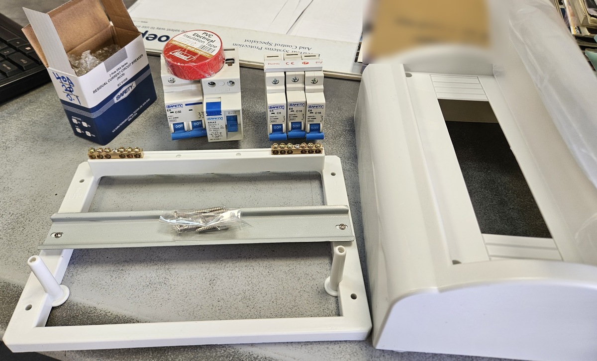 An RCCB box, a red tape roll, blue-handled circuit breakers and a white plastic distribution box base and cover on a grey desk
