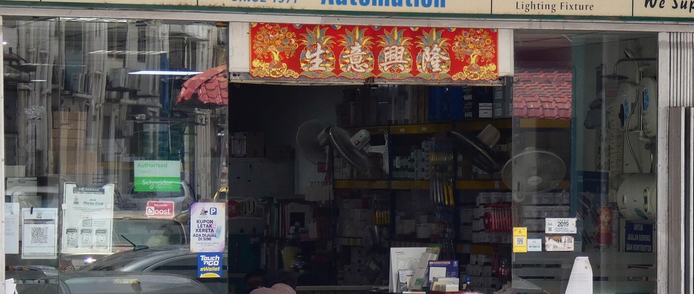 Glass shopfront with a green Schneider Electric Authorised Dealer sticker, shelves of small boxes and hanging screwdrivers, wall fans and water heaters on the right