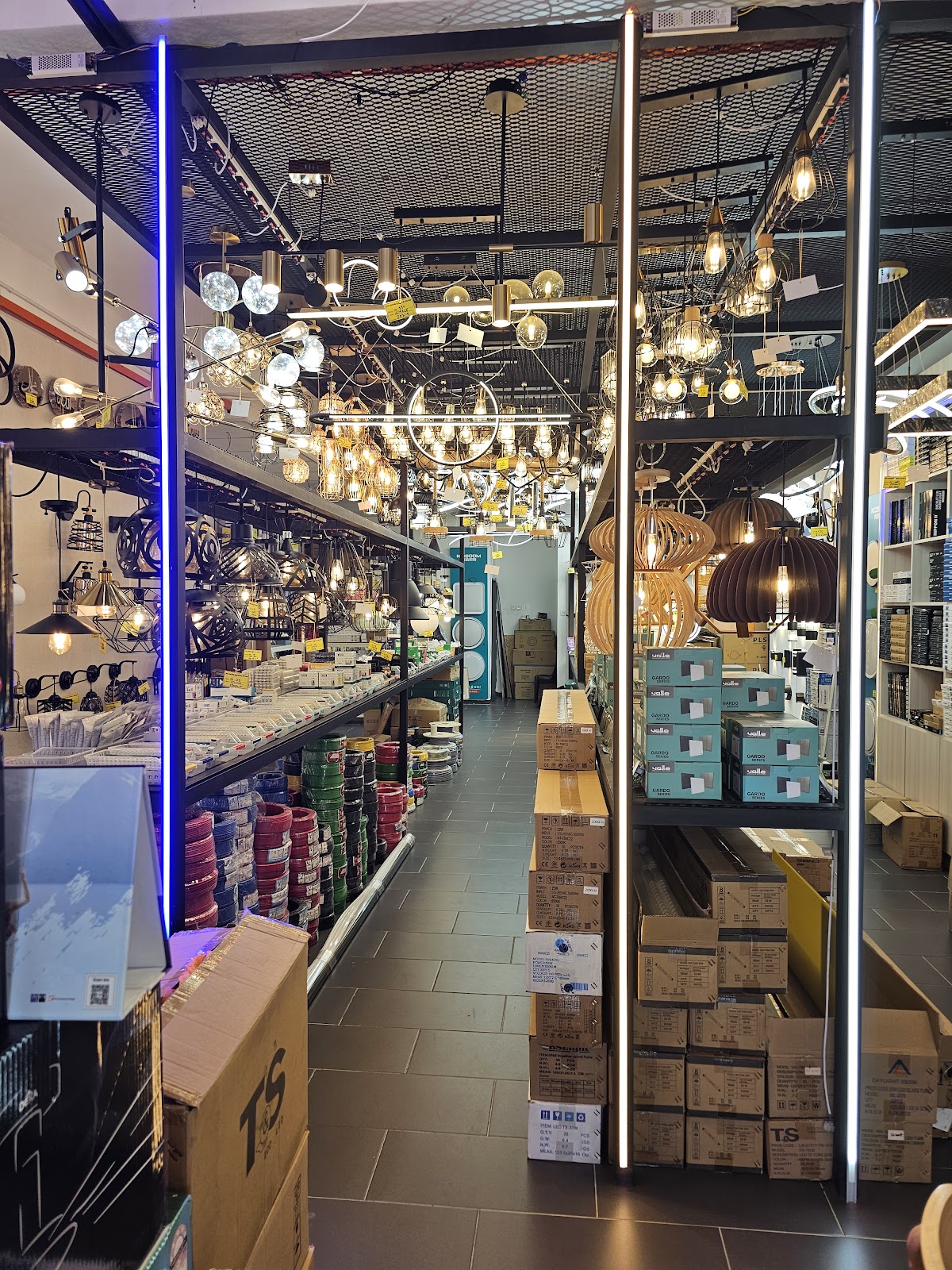 Shop aisle with rows of hanging pendant lamps above shelves of coloured wire coils and stacked switch boxes