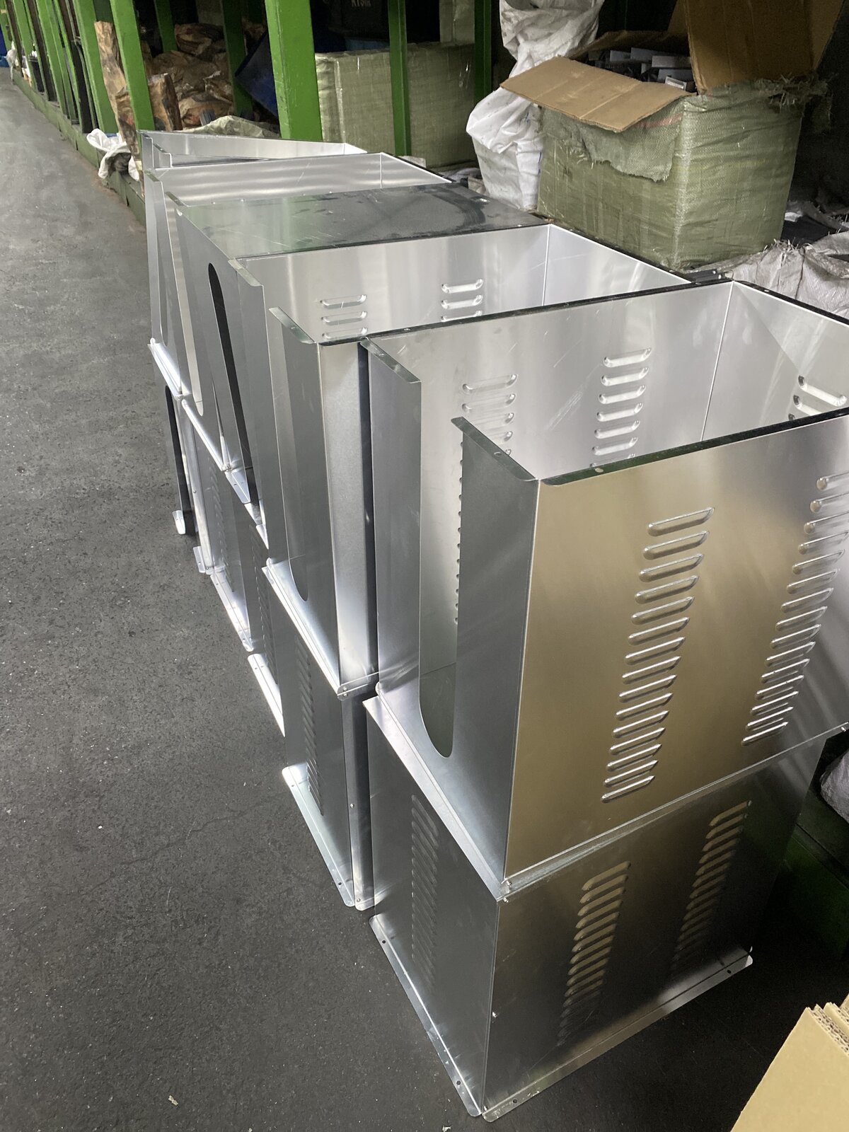 Row of bare sheet-metal boxes with louvre vents stacked two high in a warehouse aisle beside green shelving