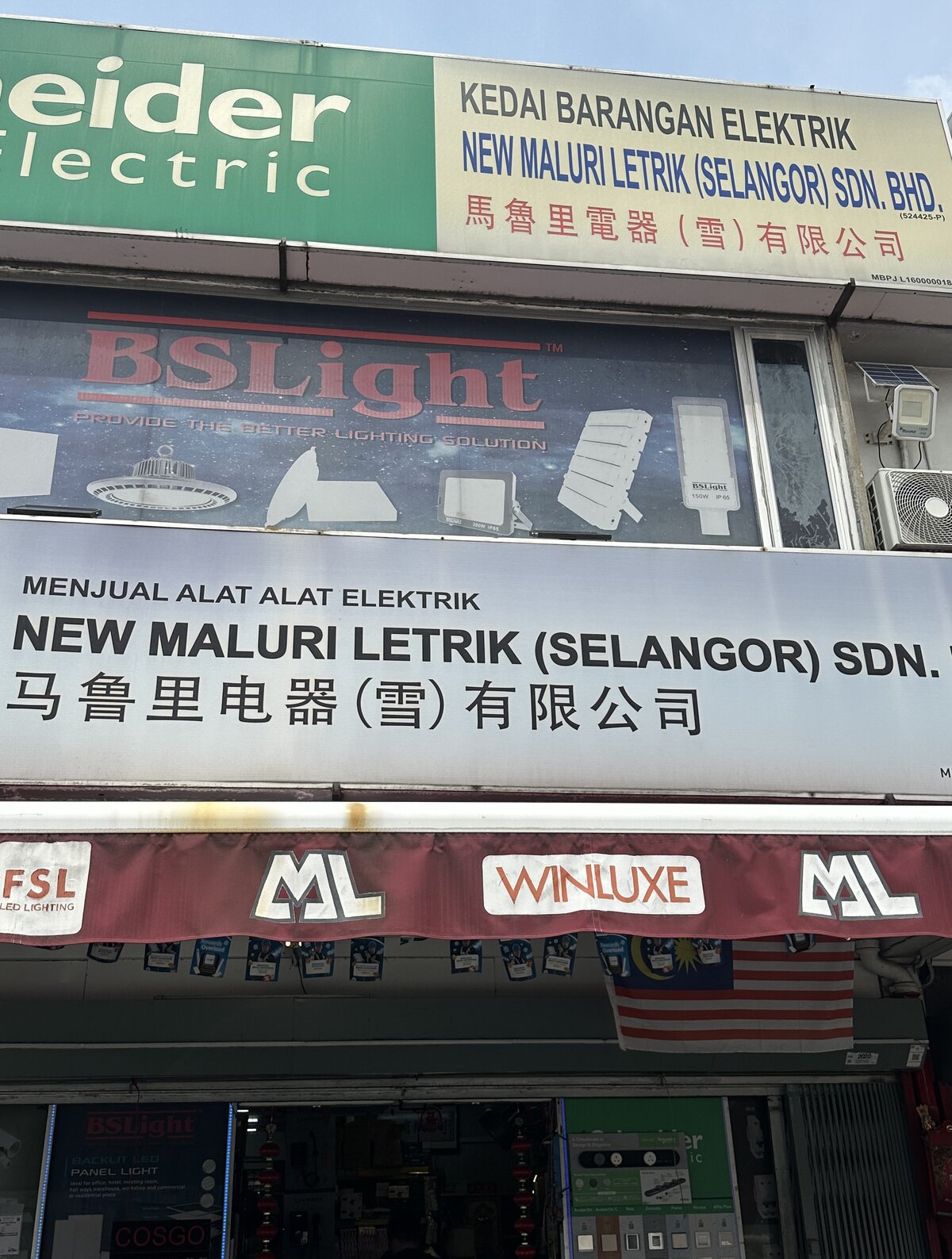 Two-storey shopfront with green Schneider Electric and white New Maluri Letrik signboards, a BSLight lighting panel, a maroon awning and switch displays at the entrance