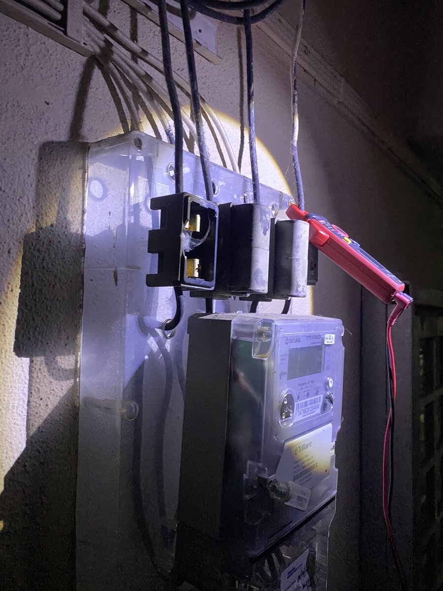 Meter board lit by a torch at night, with black service fuse holders, a digital electricity meter and a red clamp meter on one cable