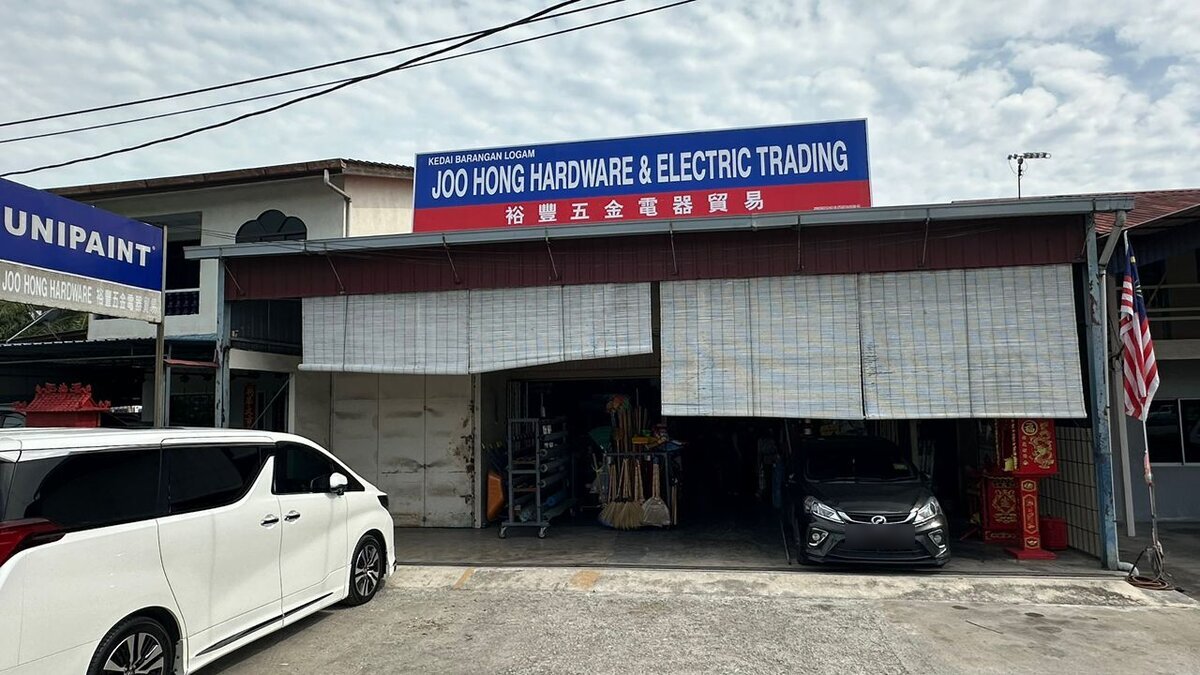 Single-storey shop with a blue and red Joo Hong Hardware & Electric Trading signboard, bamboo blinds over the front and cars parked outside