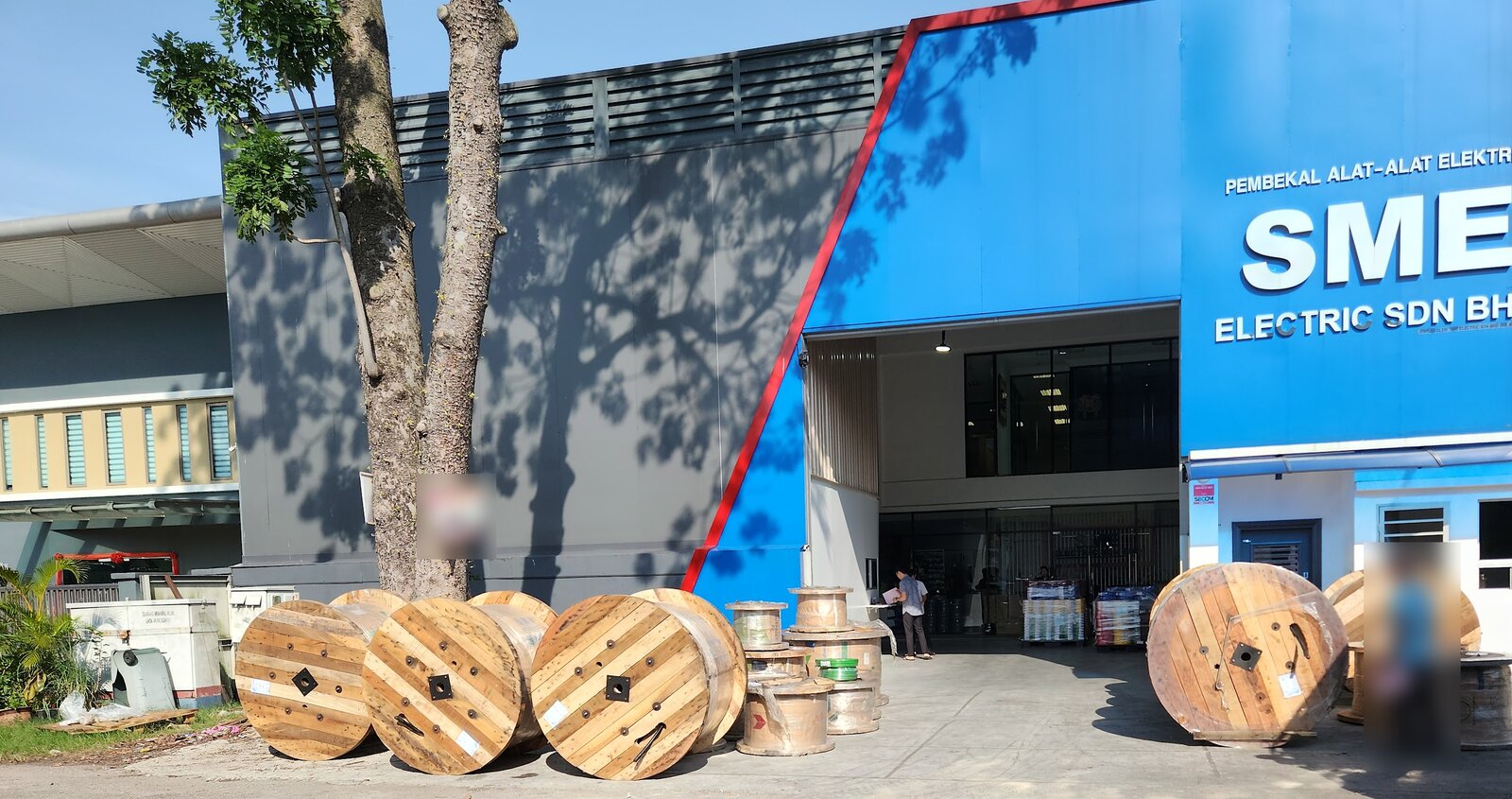 Large wooden cable drums lined up on the forecourt of a blue industrial building signed SME Electric Sdn Bhd, with pallets of goods inside the entrance
