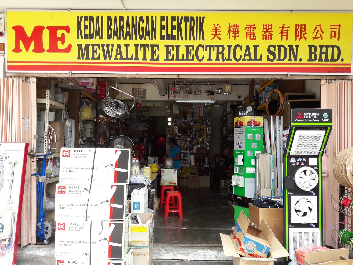 Yellow and red signboard reading Mewalite Electrical Sdn Bhd above an open shop with fan cartons, a green switch display stand and a black ventilation-fan stand