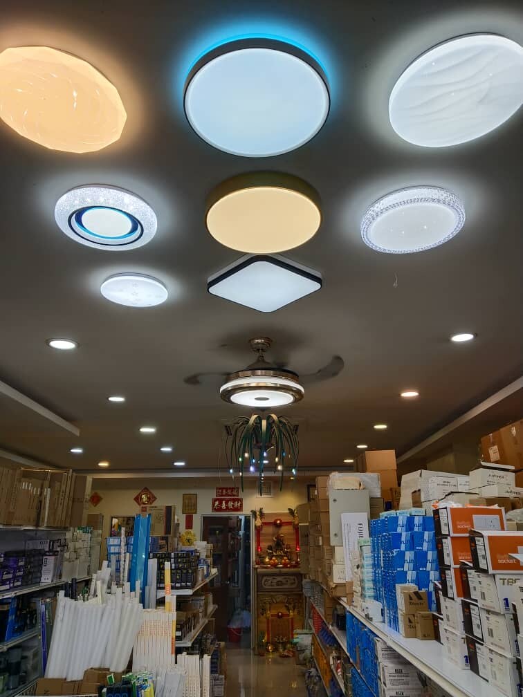 Narrow shop aisle with bundles of white LED tubes on the left, shelves stacked with blue and orange product boxes on the right, and a ceiling fan with a built-in light overhead