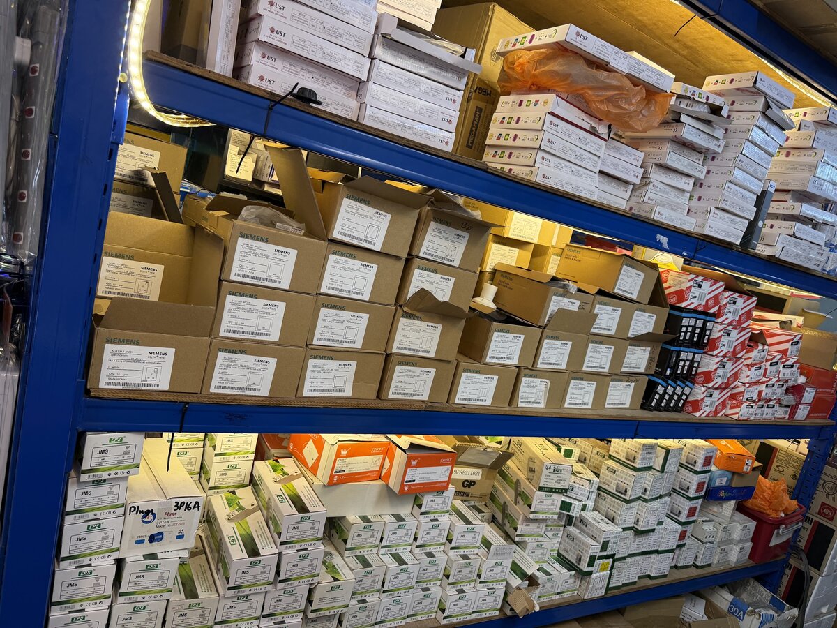 Blue steel shelving stacked with white UST lighting boxes, brown Siemens cartons, red-and-white MK boxes and many green-and-white EPS circuit-breaker boxes