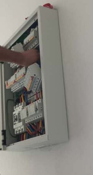 Open white wall-mounted distribution board seen from the side, with rows of white modular breakers, red, yellow and blue cables and a hand reaching inside