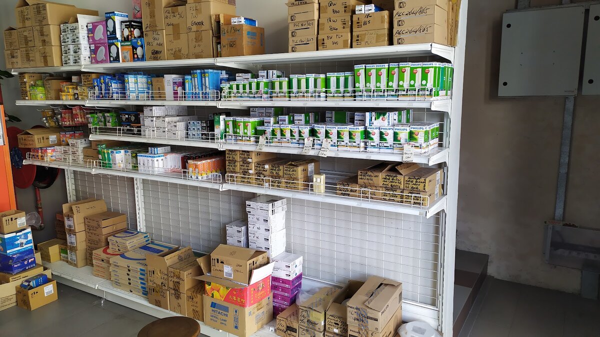 Retail shelving stacked with green and white Philips lamp boxes, cardboard cartons labelled with wattages and colour temperatures, and Hitachi spiral lamp boxes on the floor