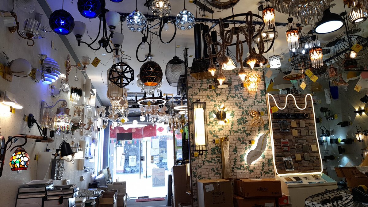 Narrow lighting showroom with pendant lamps and chandeliers hanging from the ceiling, wall lights on both sides and a lit switch-plate display stand