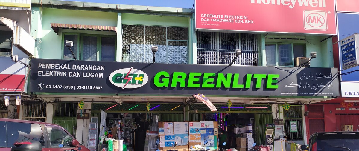 Two-unit shopfront with a black fascia reading Greenlite in large green letters, a GEH logo and two phone numbers, a red Honeywell and MK sign above, and boxed fans and ladders at the open shutters