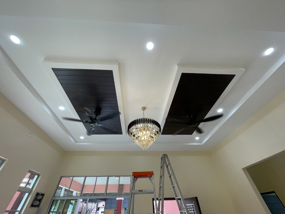 Living room with a stepped white false ceiling, recessed downlights, two dark ceiling panels with fans and a crystal chandelier, and a stepladder by the window