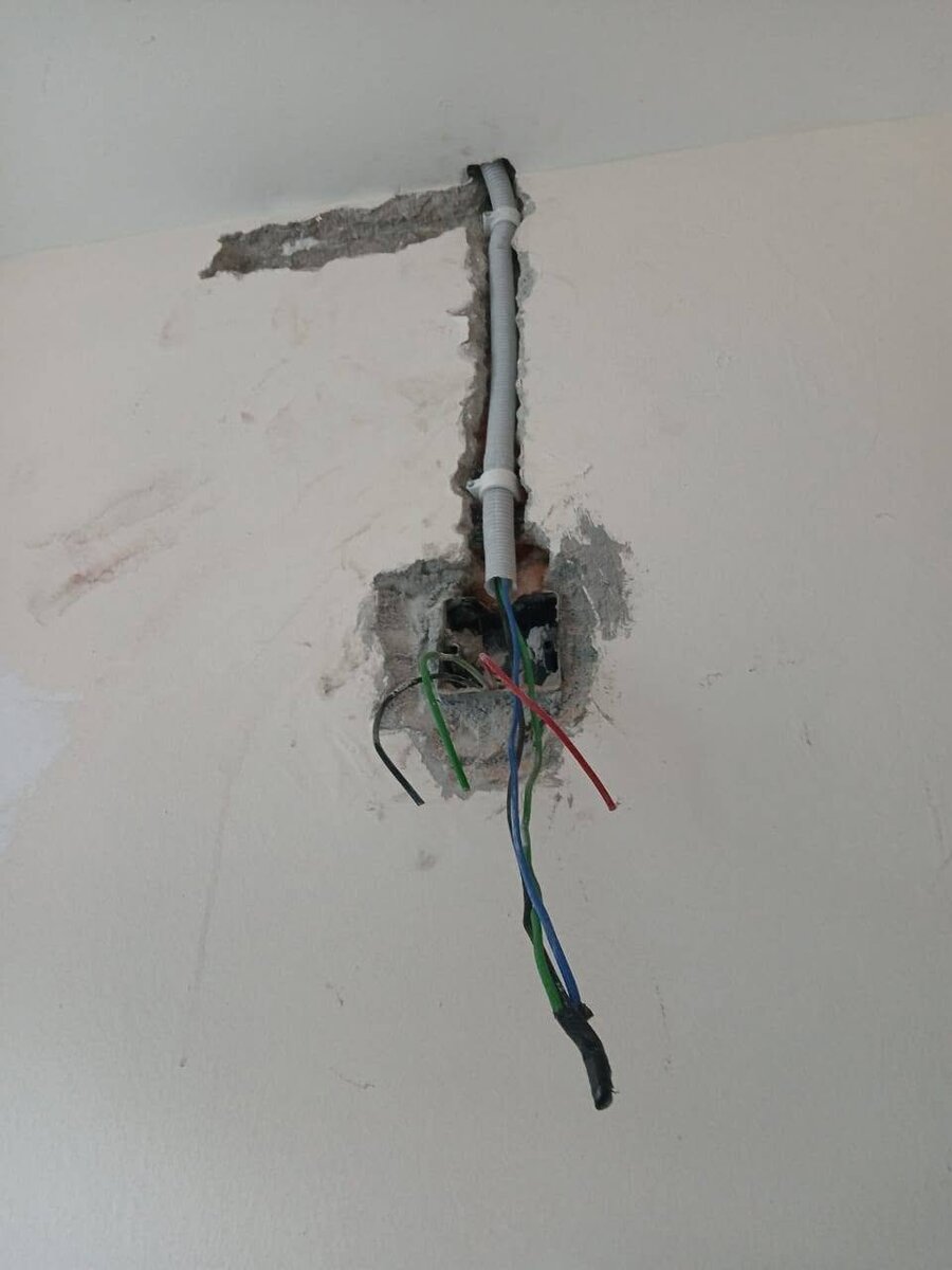 White wall with a fresh vertical groove holding a grey conduit that runs from the ceiling to a recessed box, coloured cables hanging out of the box