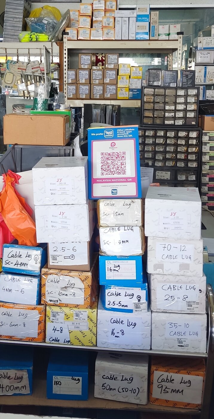 Trolley stacked with white, blue and orange boxes, most with handwritten labels such as Cable Lug SC-4mm, 25-8, 35-10, 50mm and 95mm, in front of shelves of boxed parts and parts drawers