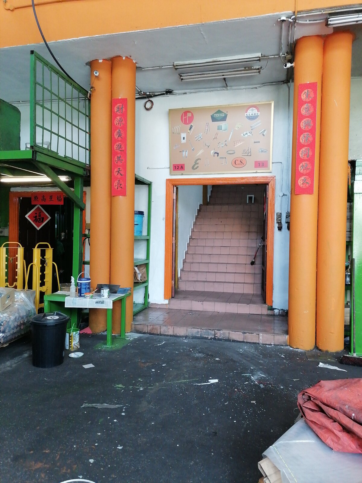 Tiled staircase entrance between two pairs of orange pillars with red Chinese couplets, under a framed board showing TOYO and GLOBE logos, product pictures and the unit numbers 12A and 13