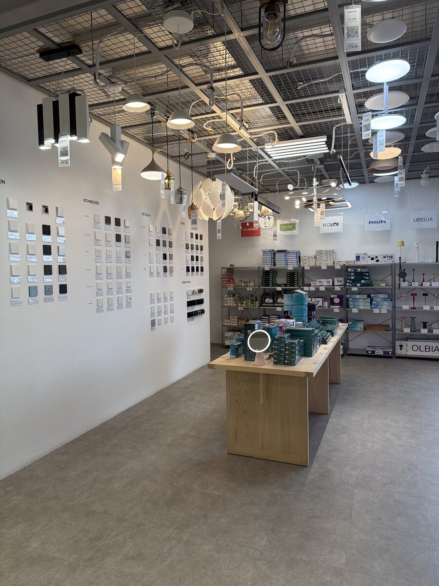 Bright showroom with rows of wall switch plates mounted on a white wall, pendant lamps hanging from a mesh ceiling, a wooden display table and metal shelves of boxed lighting products