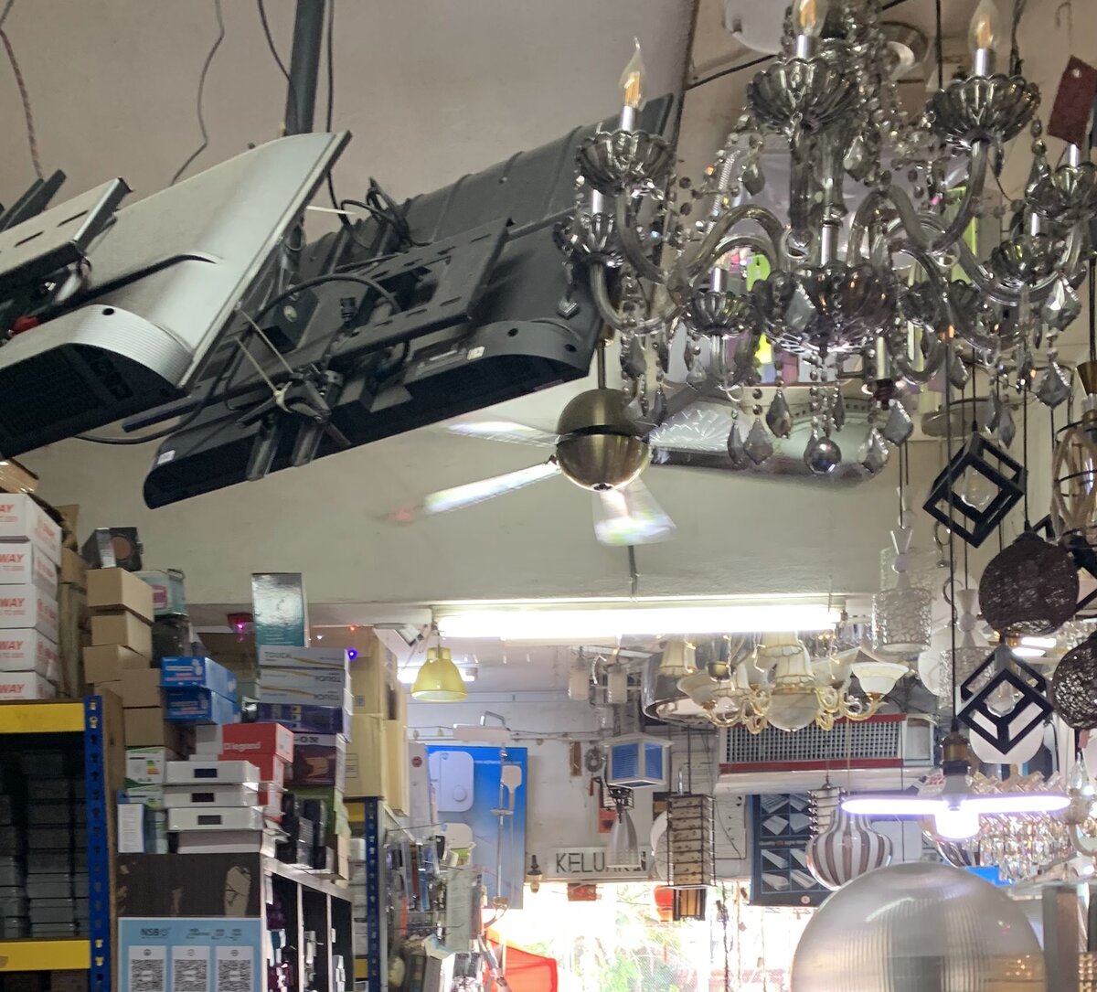 Crowded shop interior with crystal chandeliers and pendant lamps hanging from the ceiling above blue metal shelves stacked with boxed electrical goods